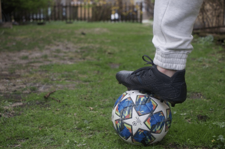 A person's foot, clad in a black shoe, rests atop a soccer ball in a green field.